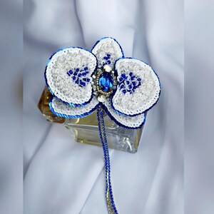 May include: A handcrafted orchid brooch featuring white and royal blue sequins, clear beads, and a central oval blue crystal. A long, dangling chain of blue and silver beads extends from the center, creating a detailed floral accessory.
