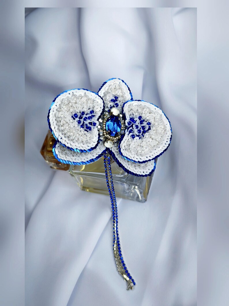 May include: A handcrafted orchid brooch featuring white and royal blue sequins, clear beads, and a central oval blue crystal. A long, dangling chain of blue and silver beads extends from the center, creating a detailed floral accessory.