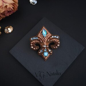 Op de afbeelding: Een fleur-de-lis broche, ingewikkeld geborduurd met bruine, blauwe en heldere kristallen. De broche heeft een centrale aquamarijnkleurige steen en is geplaatst op een donker vierkant oppervlak. Het ontwerp bevat de tekst "V.G. Natalia".