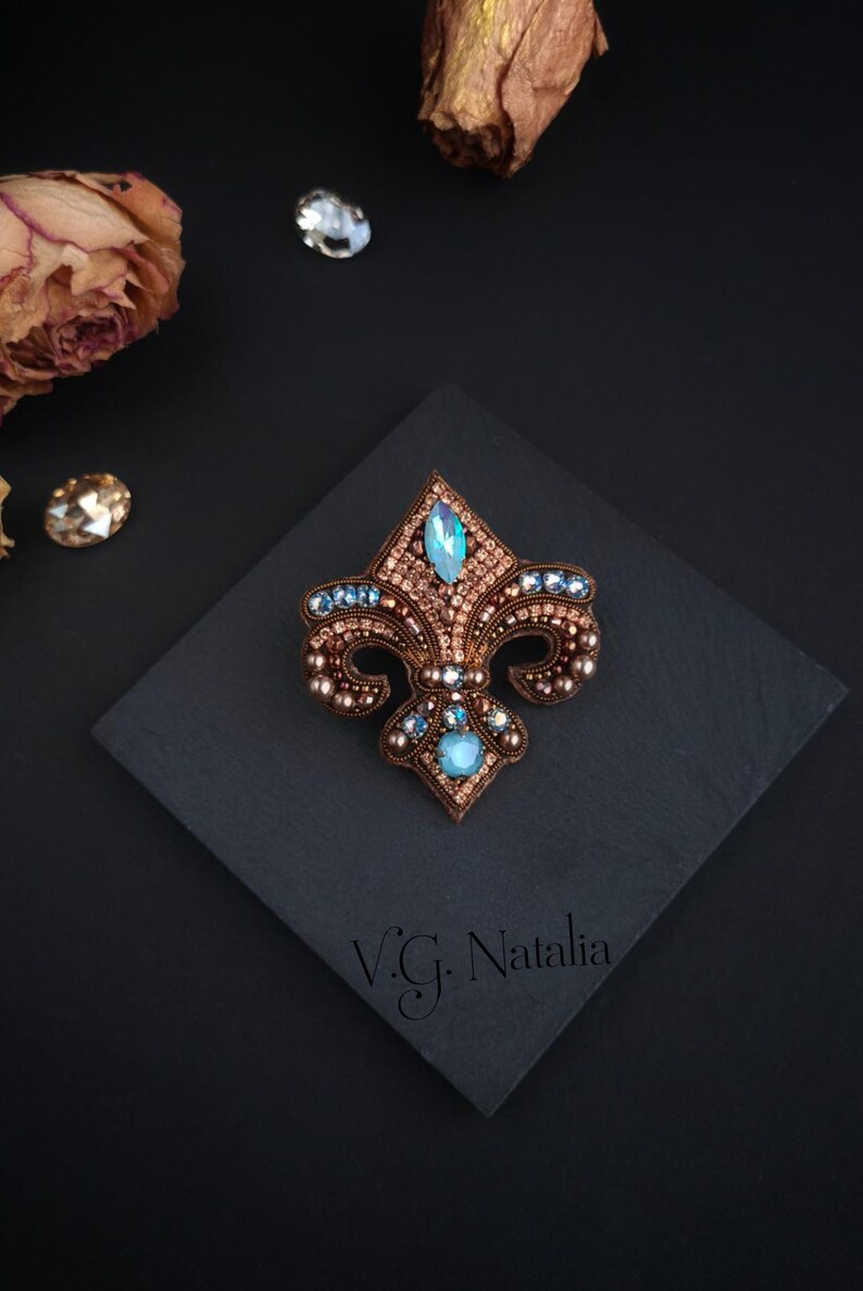 Op de afbeelding: Een fleur-de-lis broche, ingewikkeld geborduurd met bruine, blauwe en heldere kristallen. De broche heeft een centrale aquamarijnkleurige steen en is geplaatst op een donker vierkant oppervlak. Het ontwerp bevat de tekst "V.G. Natalia".