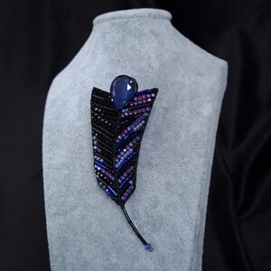 May include: A black beaded feather brooch with a blue gemstone accent. The brooch is displayed on a gray velvet stand.