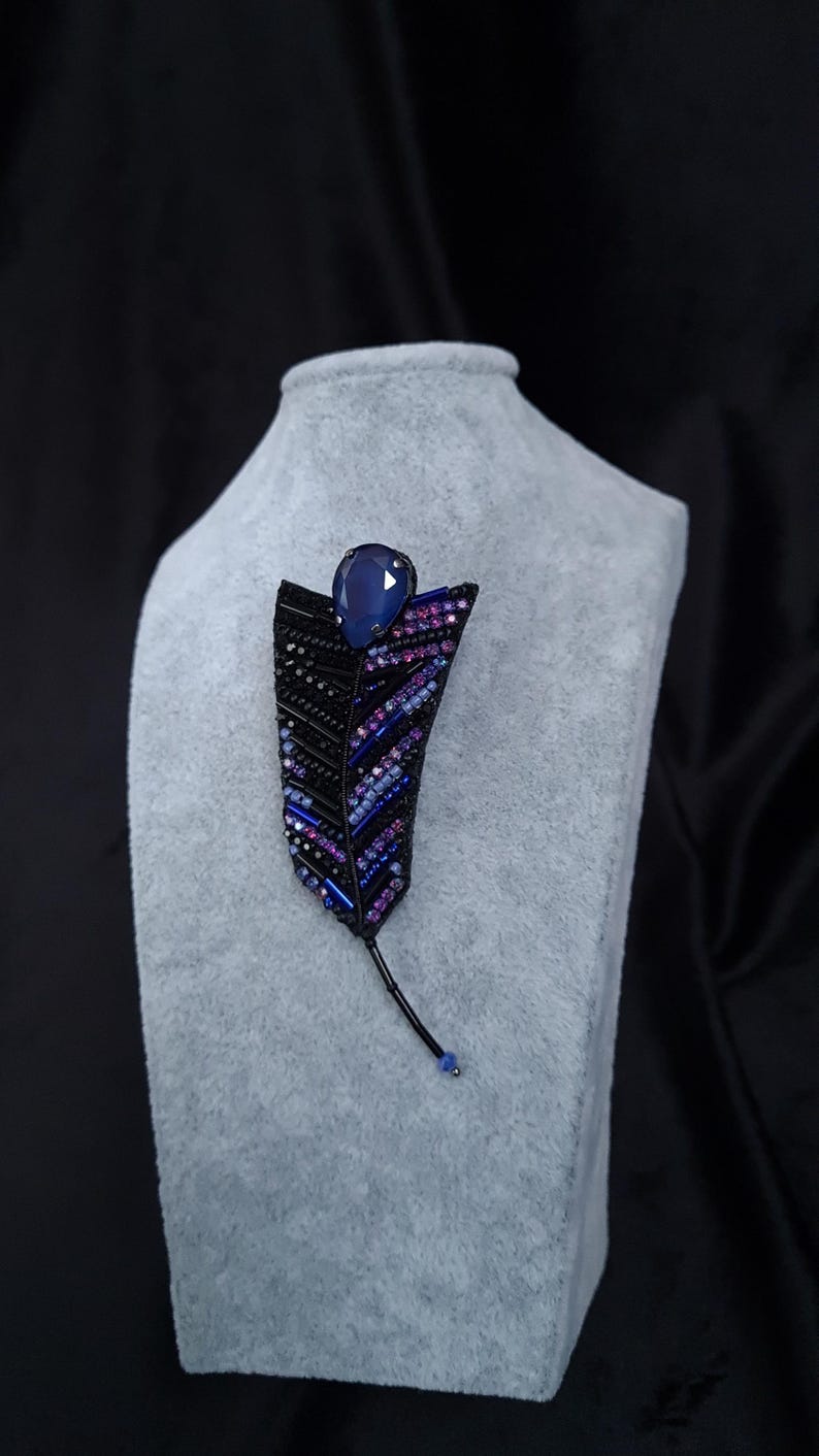 May include: A black beaded feather brooch with a blue gemstone accent. The brooch is displayed on a gray velvet stand.