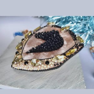 Op de afbeelding: Handgemaakte hartvormige broche met een zachte roze fluwelen basis. De broche is versierd met zwarte kralen, goudkleurige en gele kristallen en een kralenrand. Het ontwerp is ingewikkeld en gedetailleerd, geschikt voor een vleugje elegantie.