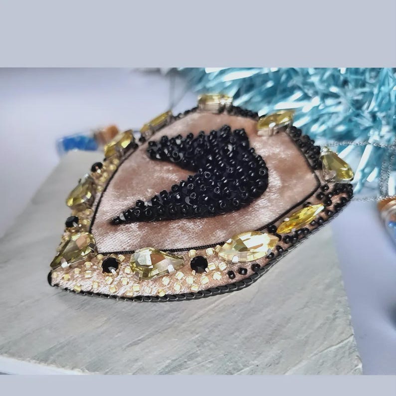 Op de afbeelding: Handgemaakte hartvormige broche met een zachte roze fluwelen basis. De broche is versierd met zwarte kralen, goudkleurige en gele kristallen en een kralenrand. Het ontwerp is ingewikkeld en gedetailleerd, geschikt voor een vleugje elegantie.