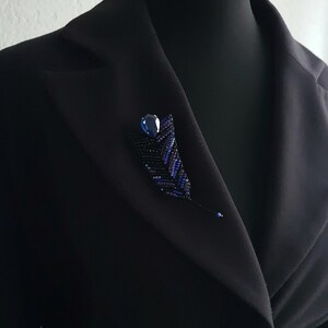 May include: A black blazer with a blue beaded brooch featuring a dark blue gemstone. The brooch is shaped like a leaf and has a delicate, intricate design.