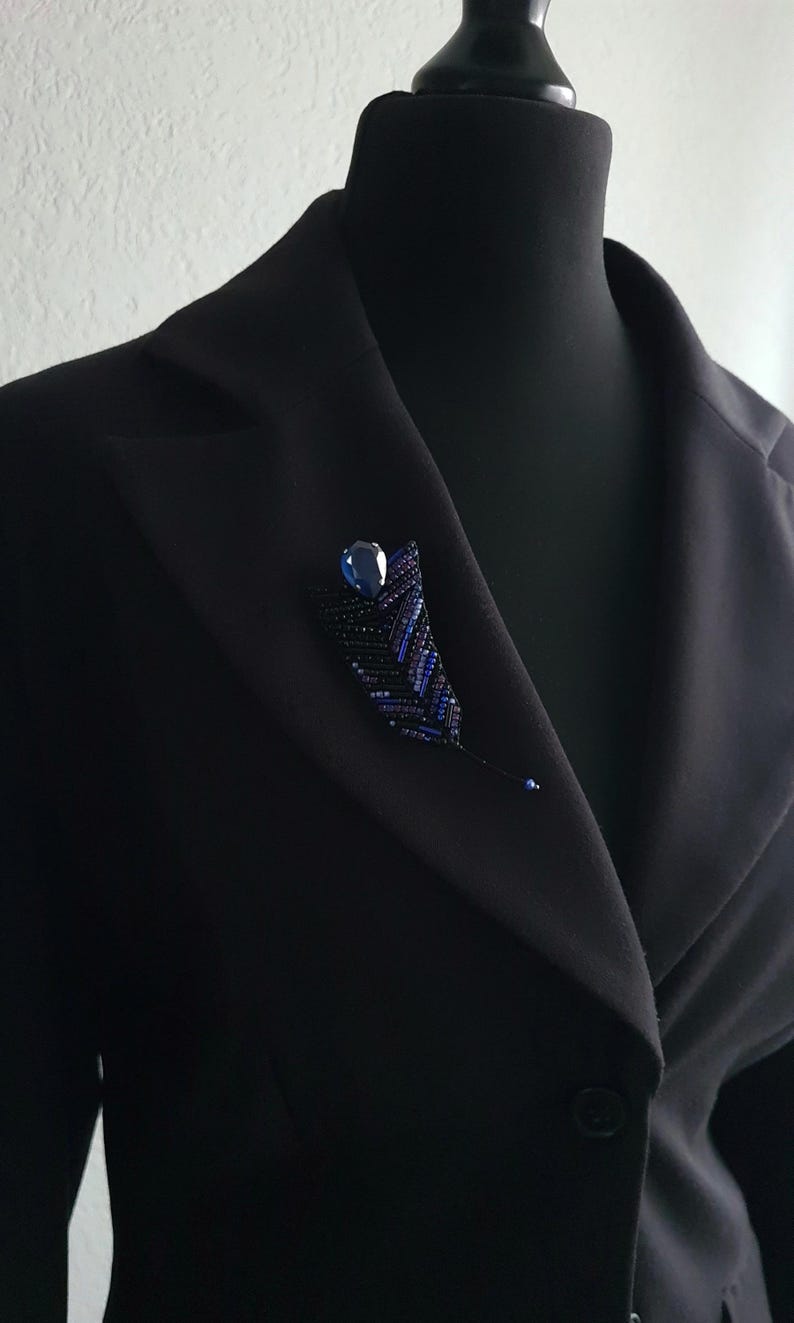 May include: A black blazer with a blue beaded brooch featuring a dark blue gemstone. The brooch is shaped like a leaf and has a delicate, intricate design.
