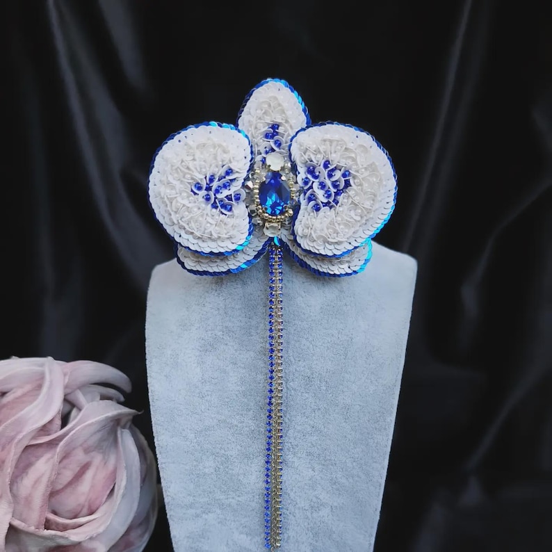 May include: A handcrafted orchid brooch featuring white and blue sequins, clear beads, and a central oval blue crystal. A long, dangling fringe of silver and blue crystal chains extends from the center, creating a detailed floral accessory.