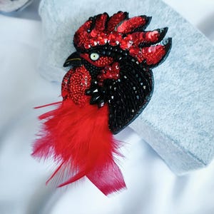 May include: A rooster brooch crafted with red and black sequins, beads, and crystals. The design features a prominent red feathered wattle, a black beak, and a single white eye with a black pupil, creating a detailed profile of a bird head.