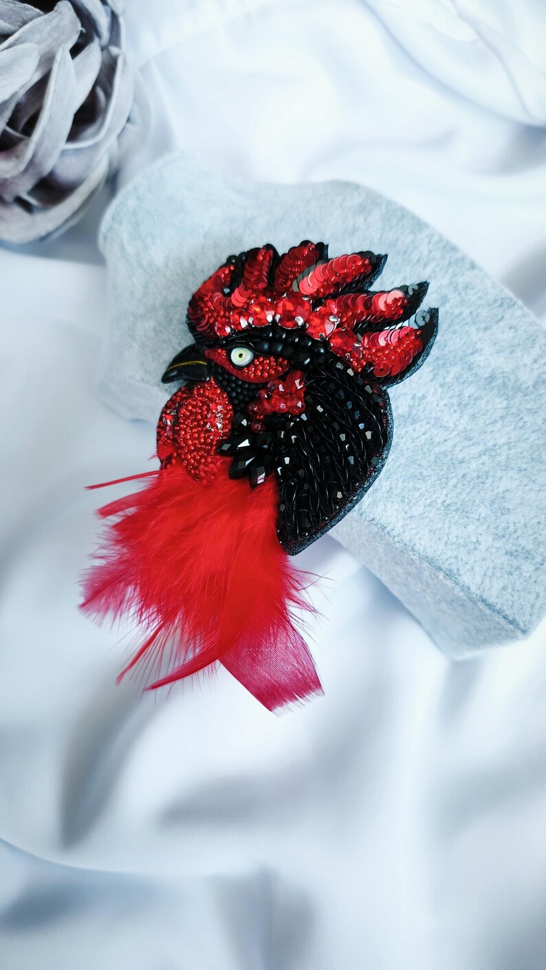 May include: A rooster brooch crafted with red and black sequins, beads, and crystals. The design features a prominent red feathered wattle, a black beak, and a single white eye with a black pupil, creating a detailed profile of a bird head.