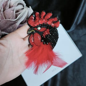 May include: A handcrafted rooster brooch featuring intricate beadwork and sequins in vibrant red and deep black. The design includes a prominent red feathered tail, a detailed crest, and a single white eye with a black pupil, creating a textured look.