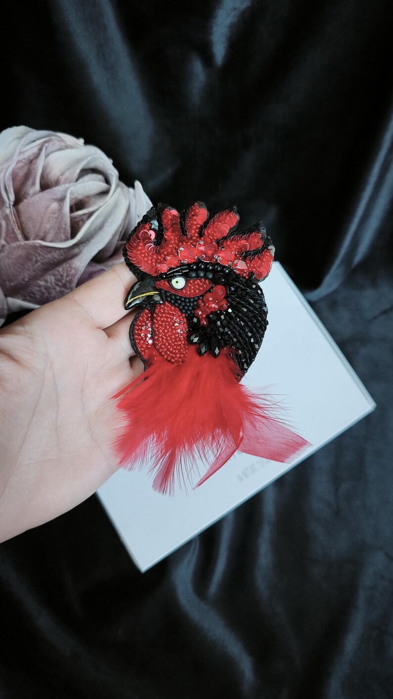 May include: A handcrafted rooster brooch featuring intricate beadwork and sequins in vibrant red and deep black. The design includes a prominent red feathered tail, a detailed crest, and a single white eye with a black pupil, creating a textured look.