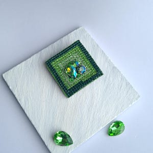 May include: A square, green brooch with a central cluster of iridescent gems. The brooch is surrounded by a darker green border and sits on a white, textured surface. Two green teardrop-shaped gems are also present.