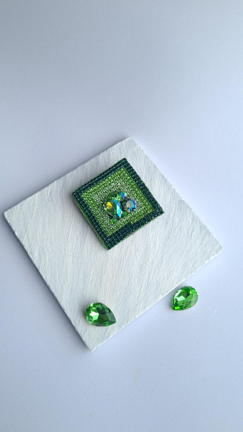 May include: A square, green brooch with a central cluster of iridescent gems. The brooch is surrounded by a darker green border and sits on a white, textured surface. Two green teardrop-shaped gems are also present.