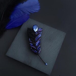 May include: A black beaded feather brooch with a blue crystal accent. The brooch is on a black surface with a blue feather in the background.
