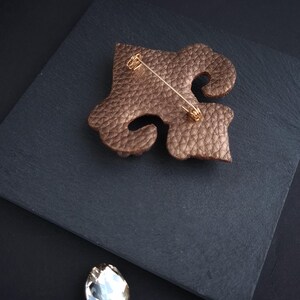 Op de afbeelding: Bronzen leren broche in een abstract ontwerp, met een goudkleurige speld. De broche ligt op een donkergrijze ondergrond, met een sprankelend kristal en een decoratief element op de achtergrond. Een elegant accessoire.