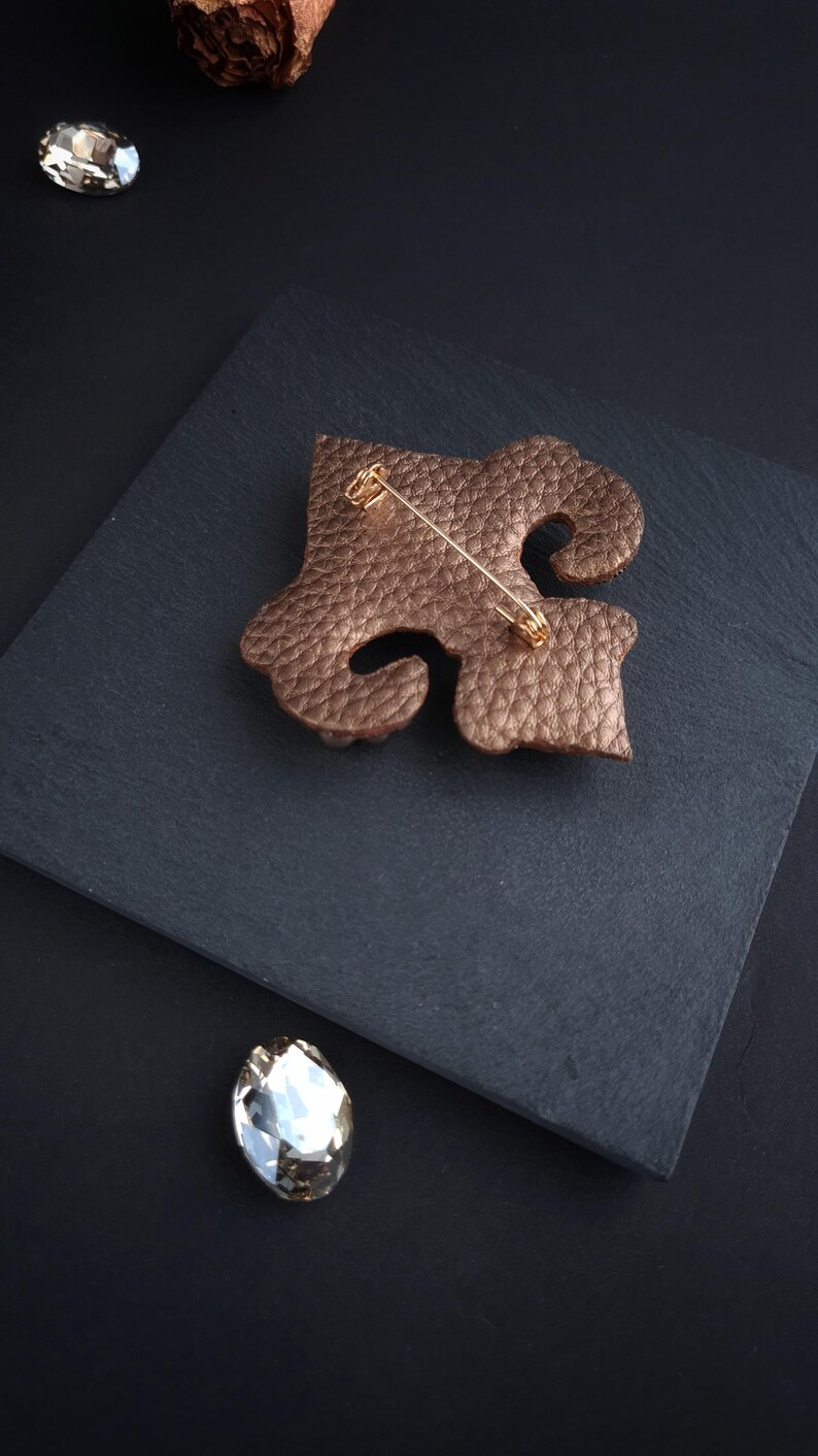 Op de afbeelding: Bronzen leren broche in een abstract ontwerp, met een goudkleurige speld. De broche ligt op een donkergrijze ondergrond, met een sprankelend kristal en een decoratief element op de achtergrond. Een elegant accessoire.
