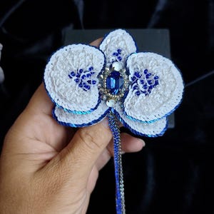 May include: A handcrafted orchid brooch featuring white and blue sequins, small white beads, and a central oval blue crystal. The flower petals are edged in deep blue, and a long, thin, dangling chain in blue and silver extends from the center.