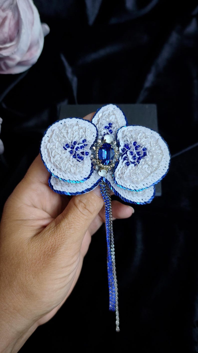 May include: A handcrafted orchid brooch featuring white and blue sequins, small white beads, and a central oval blue crystal. The flower petals are edged in deep blue, and a long, thin, dangling chain in blue and silver extends from the center.
