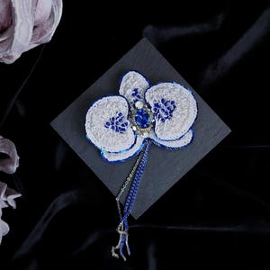 May include: A handcrafted orchid brooch featuring white and blue sequins, a central oval blue gemstone, and small blue beads. The floral design includes a dangling blue beaded chain with silver-toned accents, creating a detailed and textured accessory.