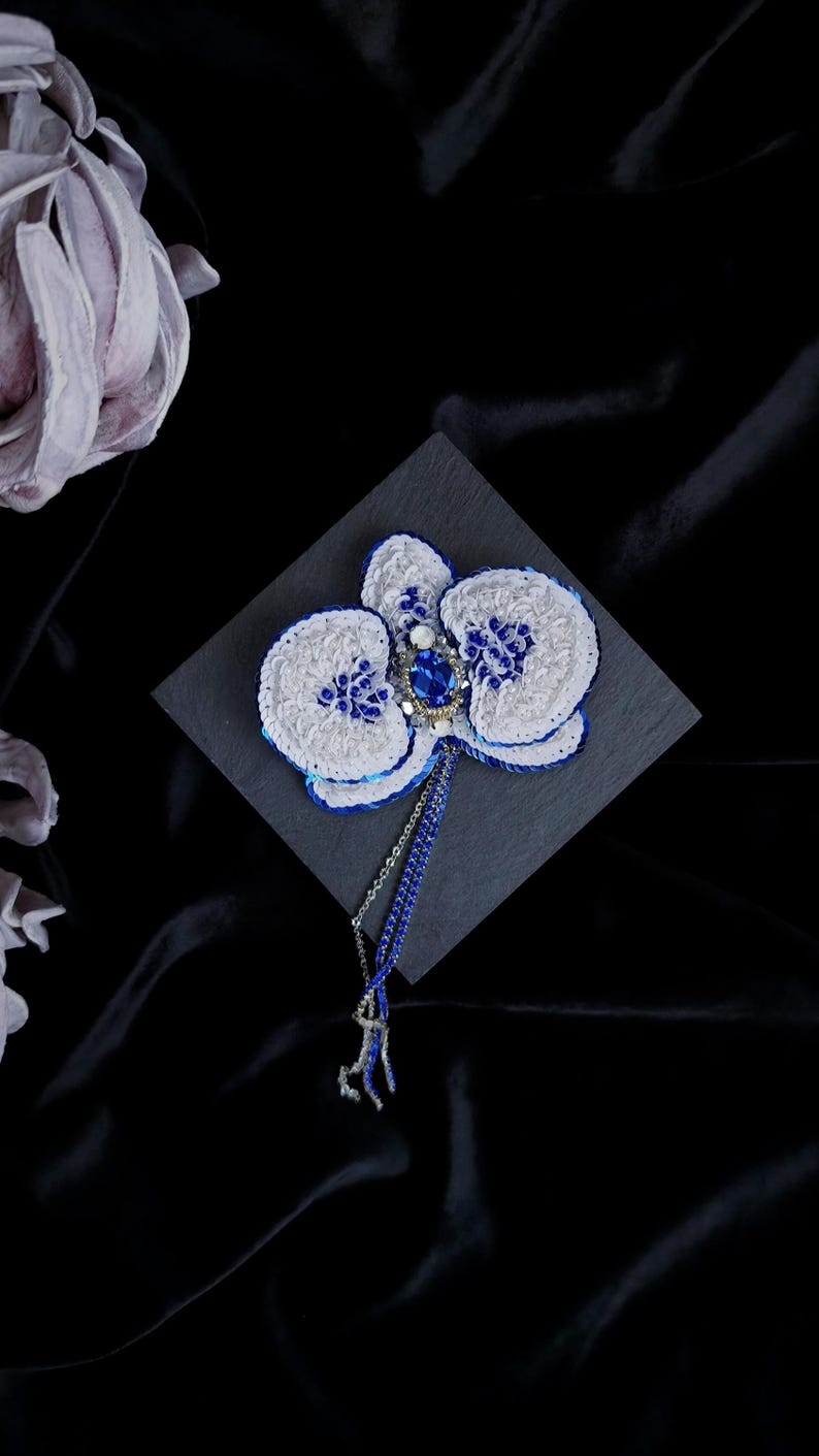 May include: A handcrafted orchid brooch featuring white and blue sequins, a central oval blue gemstone, and small blue beads. The floral design includes a dangling blue beaded chain with silver-toned accents, creating a detailed and textured accessory.