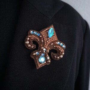 Op de afbeelding: Een gedetailleerde broche in de vorm van een fleur-de-lis. De broche heeft een bruine basis, versierd met kleine kralen en sprankelende blauwe edelstenen. Vastgemaakt op een zwarte blazer.