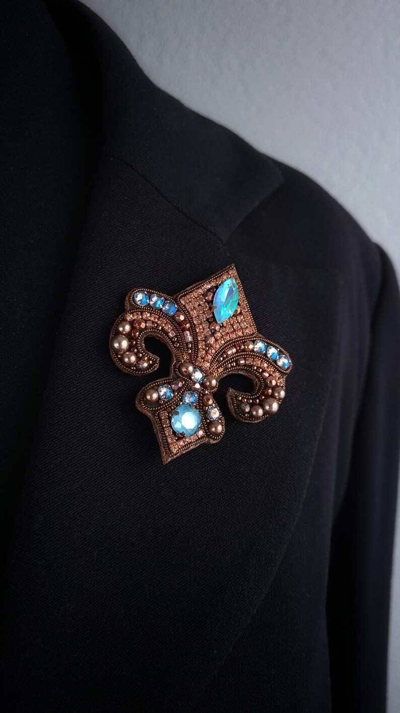 Op de afbeelding: Een gedetailleerde broche in de vorm van een fleur-de-lis. De broche heeft een bruine basis, versierd met kleine kralen en sprankelende blauwe edelstenen. Vastgemaakt op een zwarte blazer.