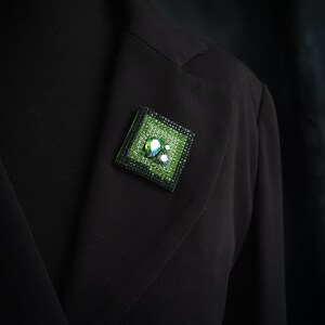 May include: A square, green brooch with a black border is pinned to a black blazer. The brooch features a textured green surface with iridescent, clear, and white embellishments. The brooch is a decorative accessory.
