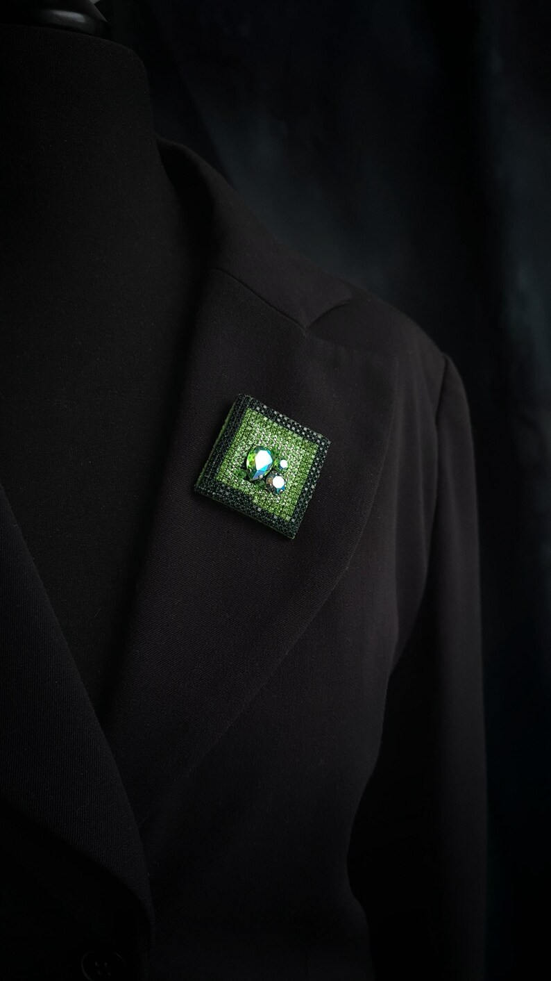 May include: A square, green brooch with a black border is pinned to a black blazer. The brooch features a textured green surface with iridescent, clear, and white embellishments. The brooch is a decorative accessory.