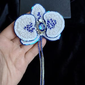 May include: A handcrafted orchid brooch featuring white and blue sequins, small white beads, and a central oval blue crystal. The flower design includes two long, dangling strands made of blue and silver rhinestones extending from the center.