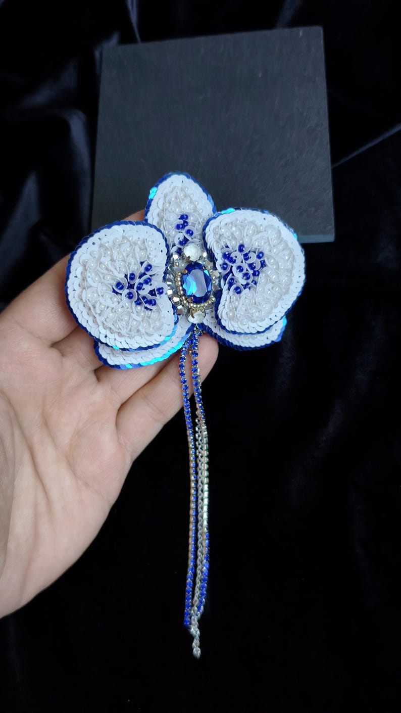 May include: A handcrafted orchid brooch featuring white and blue sequins, small white beads, and a central oval blue crystal. The flower design includes two long, dangling strands made of blue and silver rhinestones extending from the center.
