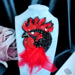 May include: A rooster brooch featuring vibrant red sequins and beads on the crest and wattle, with black beaded plumage and a sharp beak. The design includes a cluster of bright red feathers at the base, creating a textured, layered appearance.