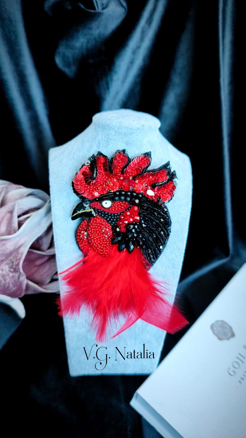 May include: A rooster brooch featuring vibrant red sequins and beads on the crest and wattle, with black beaded plumage and a sharp beak. The design includes a cluster of bright red feathers at the base, creating a textured, layered appearance.