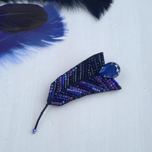May include: A beaded feather brooch with a dark blue crystal accent. The feather is made of black, purple, and blue beads.
