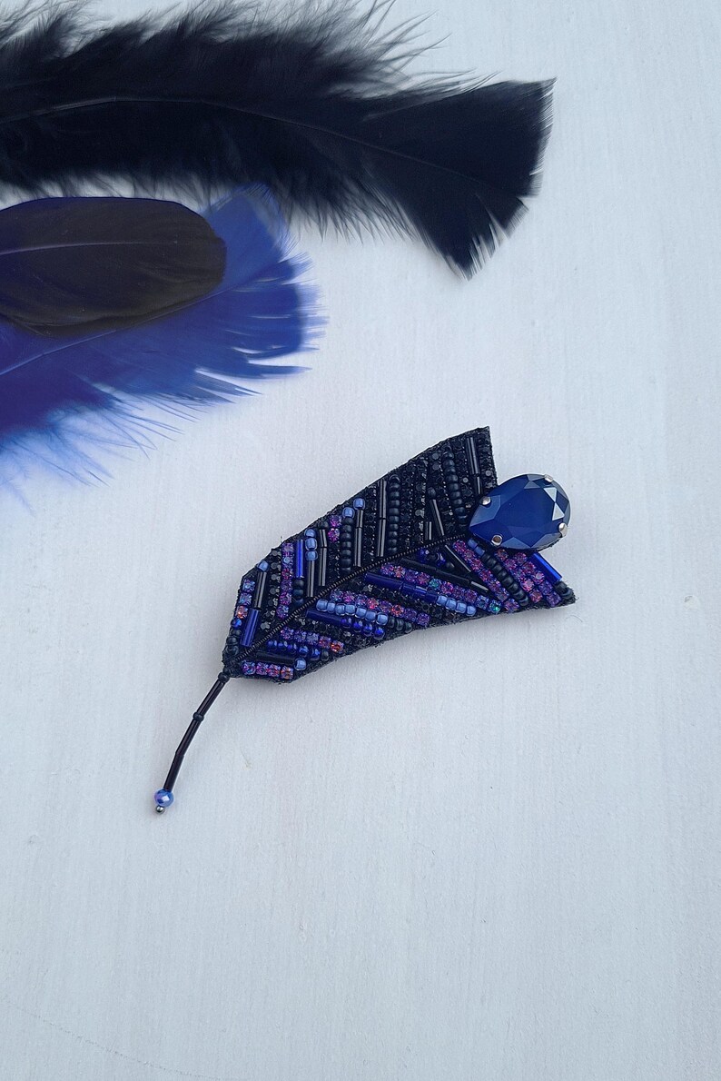 May include: A beaded feather brooch with a dark blue crystal accent. The feather is made of black, purple, and blue beads.