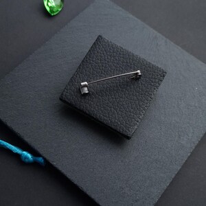 May include: A square black leather brooch with a silver pin backing. The brooch is set on a dark gray surface, with a small green gem and a blue string in the background. The leather has a textured appearance.