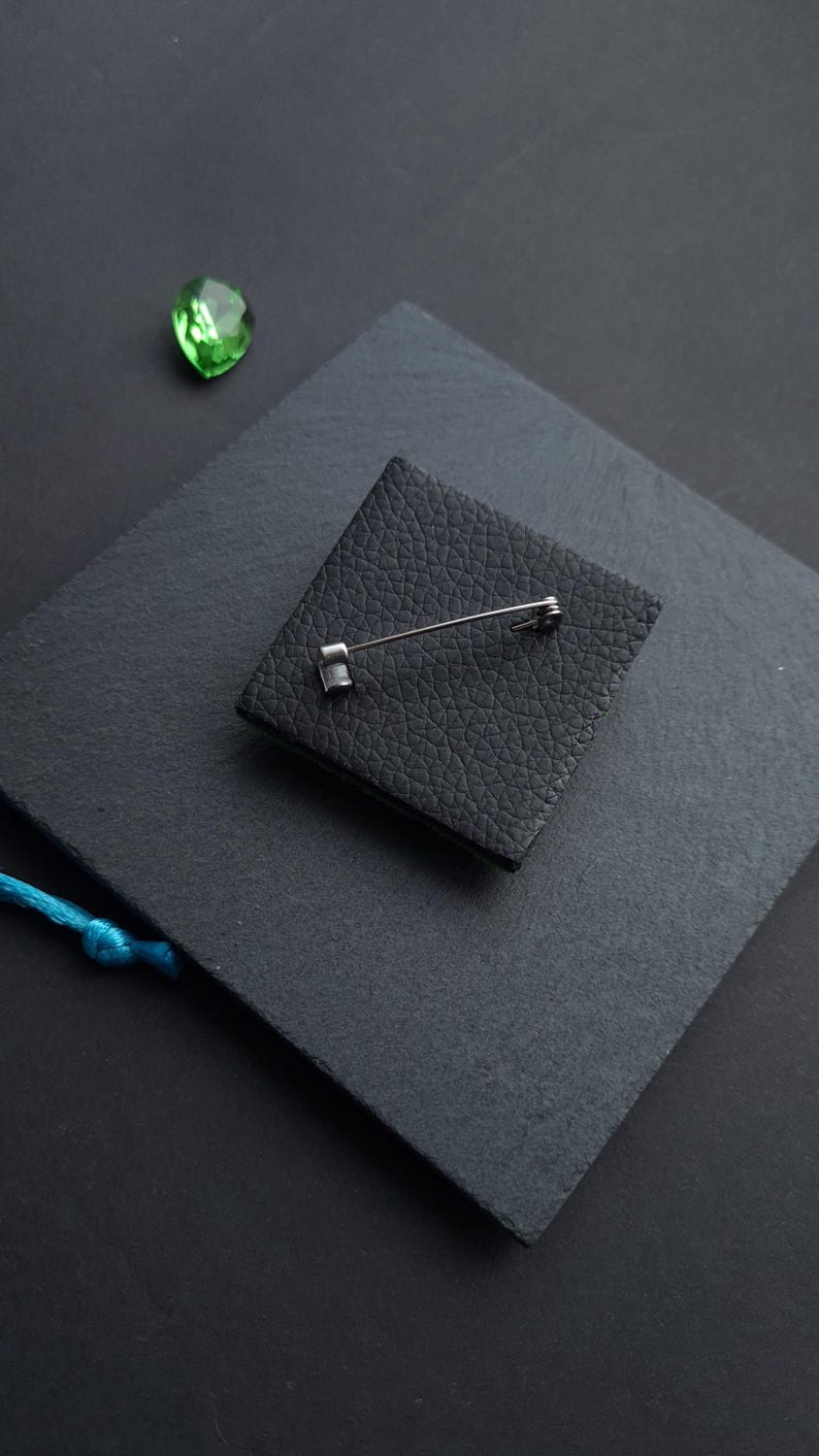 May include: A square black leather brooch with a silver pin backing. The brooch is set on a dark gray surface, with a small green gem and a blue string in the background. The leather has a textured appearance.