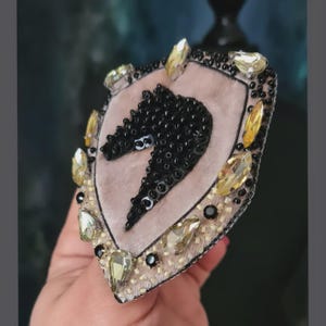Op de afbeelding: Een schildvormige broche met een zwart, met pailletten bezet paardenhoofd op een roze fluwelen achtergrond. De broche is versierd met gele en heldere kristallen en zwarte kralen. Een uniek accessoire.