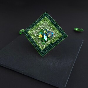 May include: A square-shaped, green brooch with a central cluster of iridescent gemstones. The brooch is embellished with smaller, sparkling green crystals and features a dark green border. Additional loose green gems are scattered around the brooch.