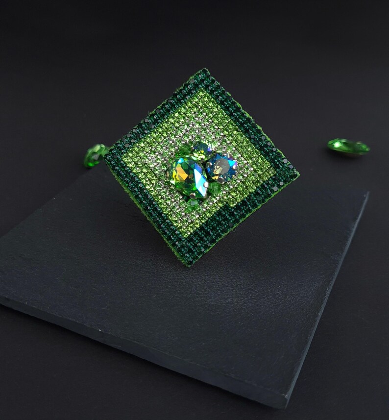May include: A square-shaped, green brooch with a central cluster of iridescent gemstones. The brooch is embellished with smaller, sparkling green crystals and features a dark green border. Additional loose green gems are scattered around the brooch.