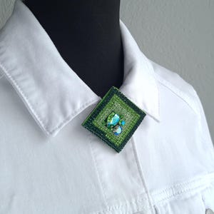 May include: A square, green brooch with a textured surface and a cluster of blue and green stones. The brooch is pinned to a white denim jacket. The jacket has a classic collar and button details.