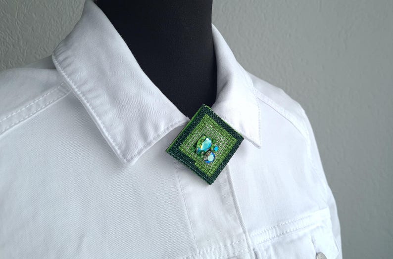 May include: A square, green brooch with a textured surface and a cluster of blue and green stones. The brooch is pinned to a white denim jacket. The jacket has a classic collar and button details.