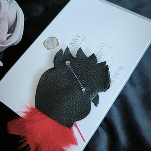 May include: A black textured brooch shaped like a rooster head with a silver metal pin on the back. The bottom of the brooch features bright red feathers. The item is resting on a white rectangular box with text that includes the words COLLECTION.