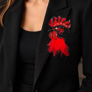 May include: A rooster-shaped brooch featuring intricate beadwork in shades of red and black, accented with sparkling sequins and a cluster of bright red feathers at the base, pinned to the lapel of a solid black tailored blazer.