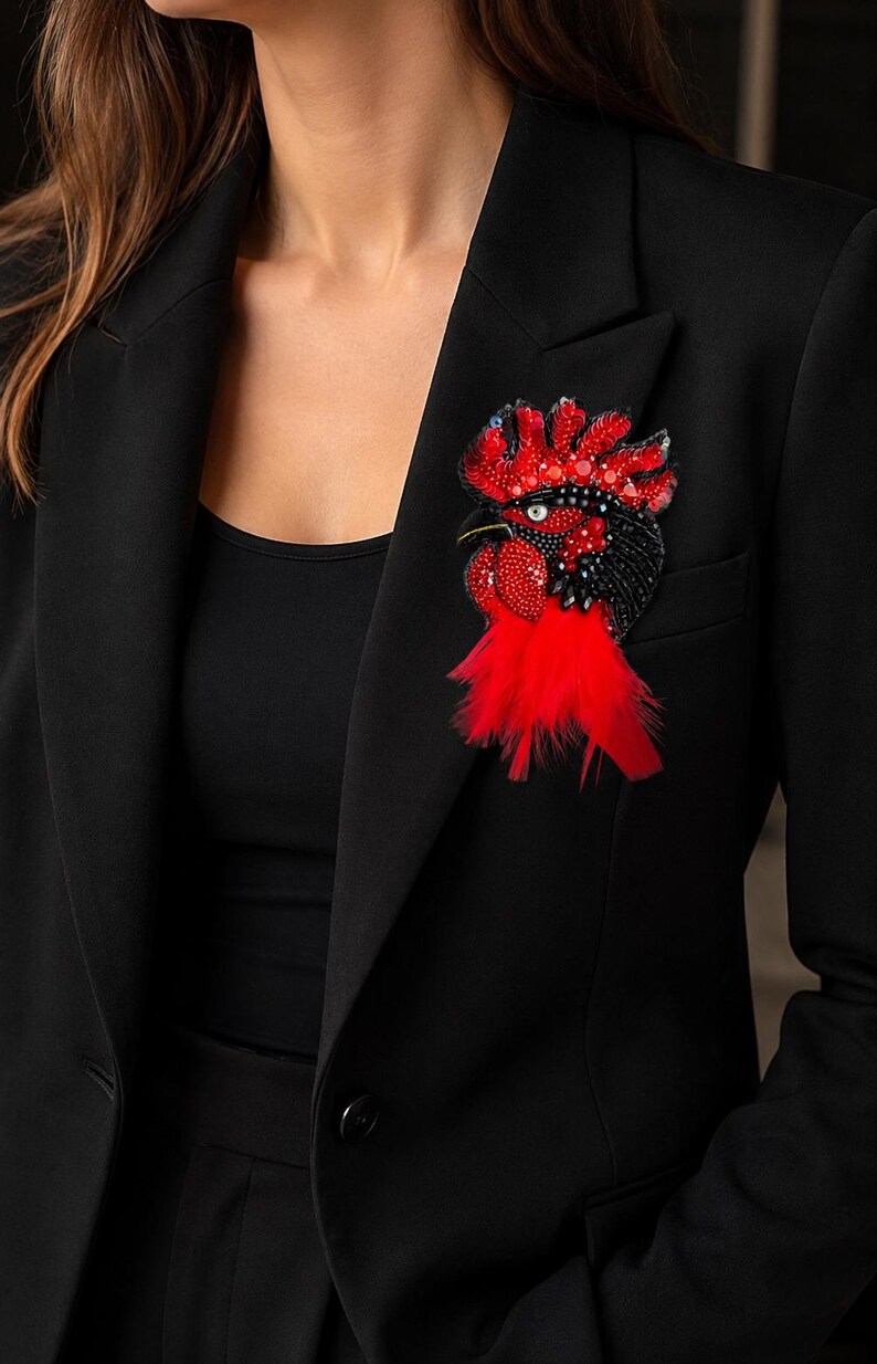 May include: A rooster-shaped brooch featuring intricate beadwork in shades of red and black, accented with sparkling sequins and a cluster of bright red feathers at the base, pinned to the lapel of a solid black tailored blazer.