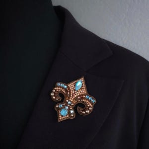 Op de afbeelding: Een fleur-de-lis broche met een bronskleurige basis, versierd met kleine, ronde kralen en grotere, lichtblauwe, druppelvormige edelstenen. De broche is vastgespeld op een zwarte blazer.