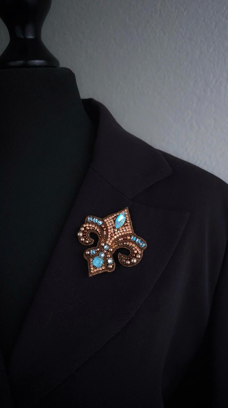 Op de afbeelding: Een fleur-de-lis broche met een bronskleurige basis, versierd met kleine, ronde kralen en grotere, lichtblauwe, druppelvormige edelstenen. De broche is vastgespeld op een zwarte blazer.