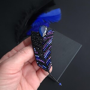 May include: A black beaded feather brooch with a dark blue rhinestone in the center. The feather is embellished with purple and blue beads.