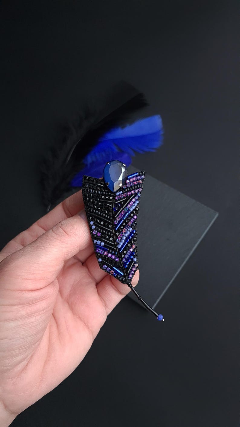 May include: A black beaded feather brooch with a dark blue rhinestone in the center. The feather is embellished with purple and blue beads.
