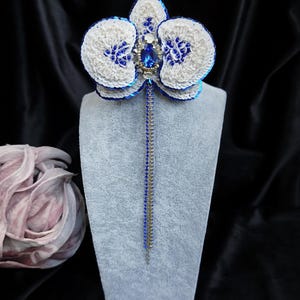 May include: A handcrafted orchid brooch featuring white and blue sequins, a central teardrop-shaped blue crystal, and small clear rhinestones. Two long, thin dangling chains, one blue and one silver, extend downward from the center of the flower.