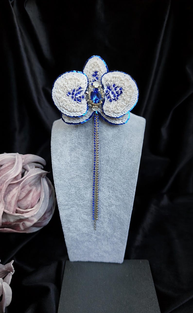 May include: A handcrafted orchid brooch featuring white and blue sequins, a central teardrop-shaped blue crystal, and small clear rhinestones. Two long, thin dangling chains, one blue and one silver, extend downward from the center of the flower.